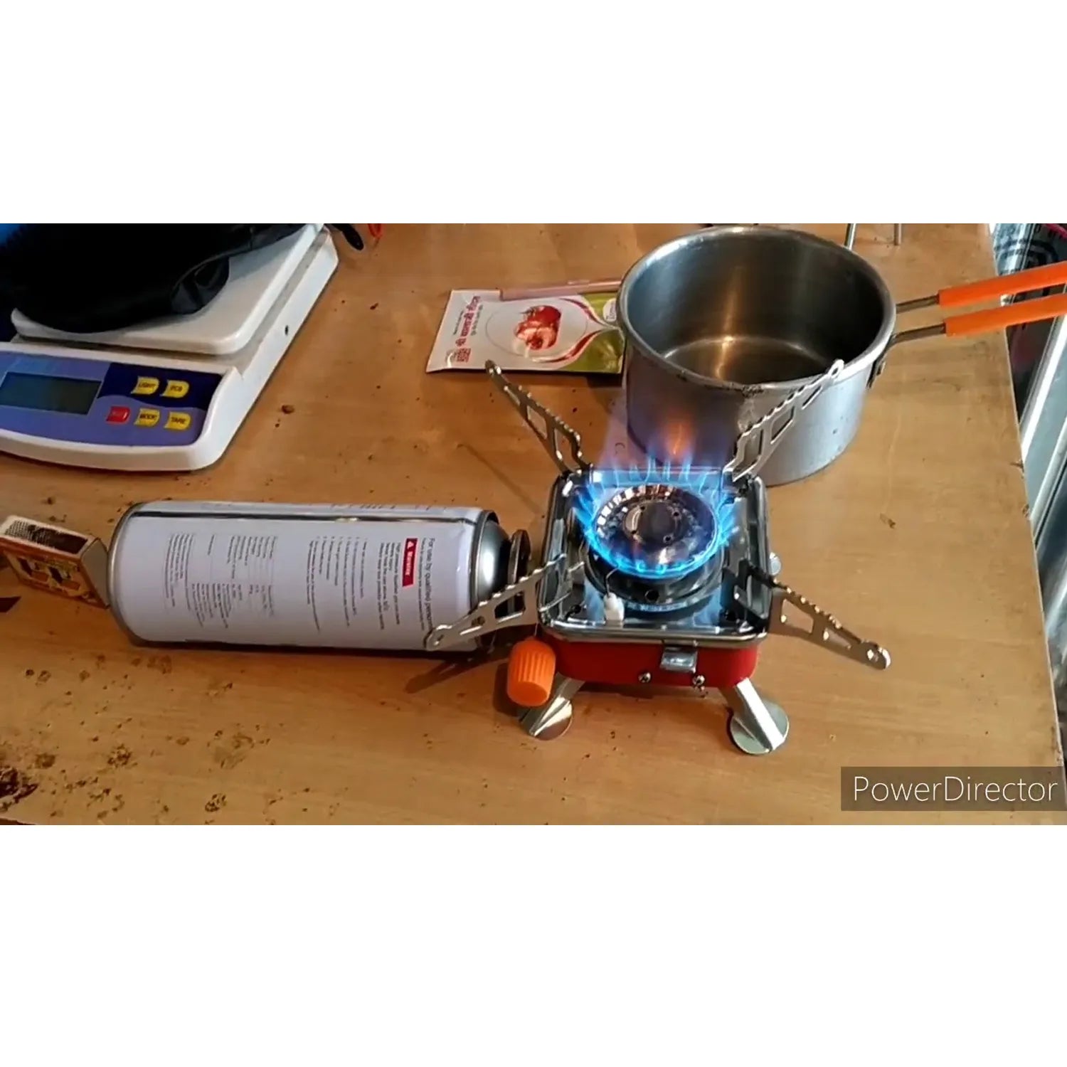 Portable gas stove with a lit burner on a wooden surface, next to a canister and a pot.